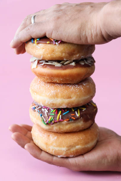 istock Close-up image of unrecognisable person sandwiching stack of alternating plain and chocolate glazed ring doughnuts between palms, fried dough dessert decorated with multicoloured sugar sprinkles or flaked almond nuts, pink background, focus on foreground 1874785116