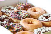 Full frame image of cardboard box of glazed ring doughnut, plain and chocolate glazed fried dough dessert decorated with multicoloured sugar sprinkles or flaked almond nuts, elevated view, focus on foreground