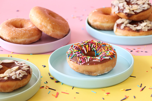 Stock photo showing close-up view of plain and chocolate glazed ring doughnuts on pastel pink and green plates. Some of the chocolate glazed cakes are topped with flaked almonds, whilst others are decorated with white, pink, green, blue, yellow, and orange, hundred and thousand sugar sprinkles.
