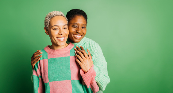 Two young cheerful women wearing green clothes embracing in a studio with a green background.