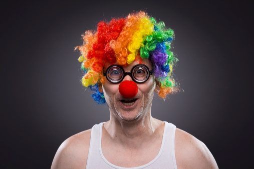 a mad evil redhead clown, wearing a white and red striped costume with a white ruff, stands with his arms on his hips and a creepy smile in front of the weathered tent of the circus