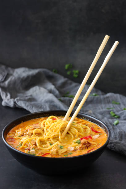 Image of wooden chopsticks sticking upright in bowl of homemade, spicy coconut milk laksa soup with chicken, noodles, chopped peanuts, spring onion and red chilli garnish, rudeness and bad luck, grey muslin, black background, focus on foreground stock photo