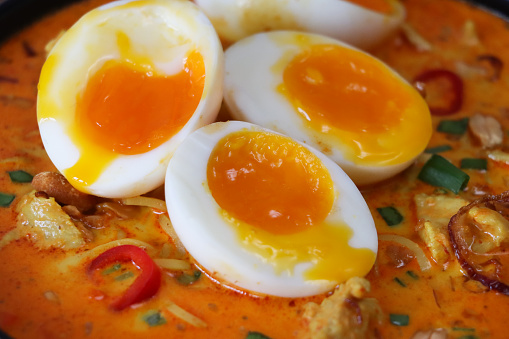 Stock photo showing close-up, elevated view of a bowl of chicken laksa soup garnished with two halved soft boiled egg, chopped peanuts, spring onion and red chilli. The main ingredients of Laksa are noodles, coconut milk and curry spice.