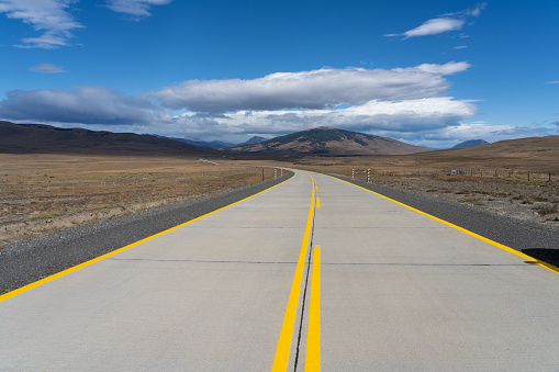 A long road with yellow lines leading towards mountains that crosses the Atacama desert in Chile. Sun with clouds in the blue sky.