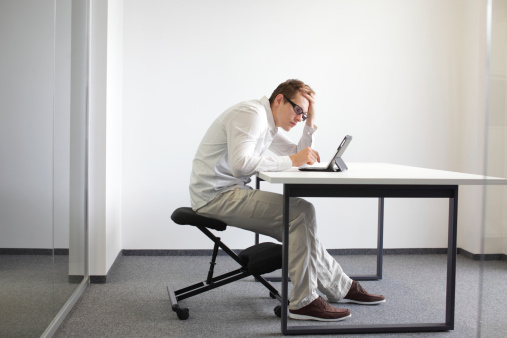Young man is bent over his tablet in his office,seating on kneeling chair Bad sitting posture at work