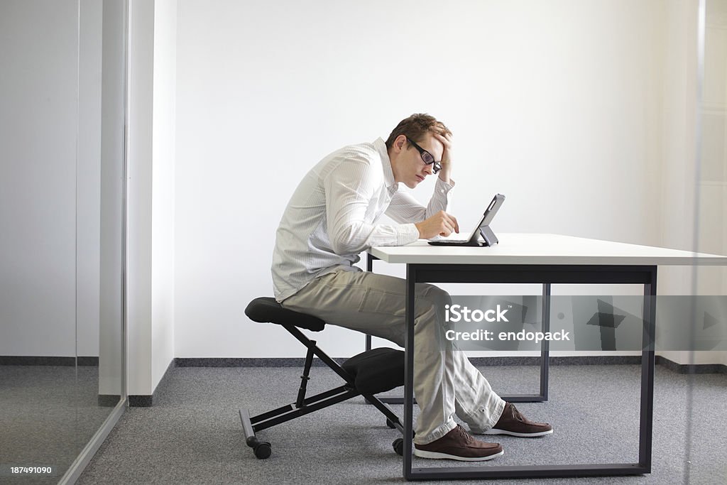man is bent over  tablet.Bad sitting posture at work Young man is bent over his tablet in his office,seating on kneeling chair Bad sitting posture at work Bad Posture Stock Photo