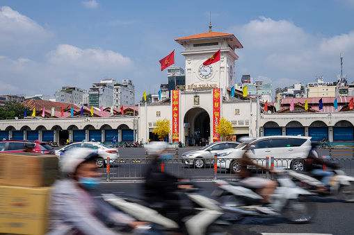 Ho Chi Minh City, Vietnam - February 14, 2023: Motorcycles and cars pass by the main entrance and clock tower of Ben Thanh Market.