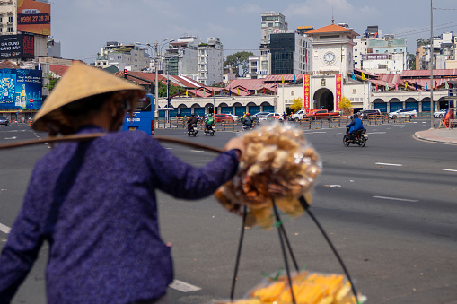 Ho Chi Minh City, Vietnam - February 14, 2023: A street vendor carrying baskets suspended from a shoulder pole walks around a traffic intersection at Ben Thanh Market.