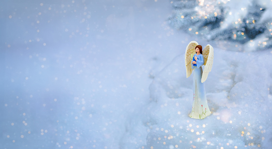 Christmas angel with book in hand in snow near Christmas tree in evening. Outdoor nature photo. Snowy weather.
