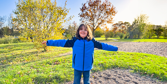 A cute little boy with Down's syndrome, his arms raised, smiling with joy and happiness as he looks into the camera. A child in a public park on a sunny winter's day, wearing a blue jacket and with dark blonde hair.
