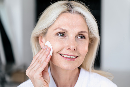 Mature woman with healthy smooth skin applying hyaluronic moisturizing toner with cotton pads at home bathroom. Daily hygiene procedures and skincare routine.