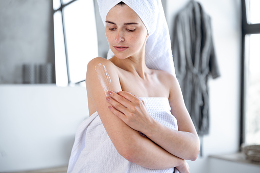 Woman wrapped in bath towel putting nourishing lotion on shoulder. Soft skin effect, enjoying natural organic cosmetic product. Beauty, skincare, bodycare concept.