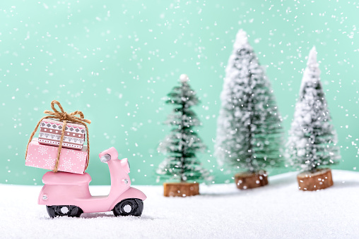 Pink toy motorbike with a Christmas gifts next to fir trees while it is snowing. Concept of celebrating Christmas and New Year.