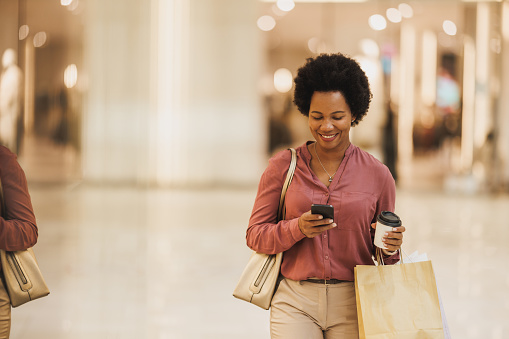 Shot of a happy Black woman using her cell phone while holding shopping bags and cup of coffee to go at the city mall.
