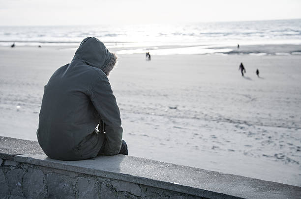 Solitary person in hooded parka coat sits on sea wall alone stock photo