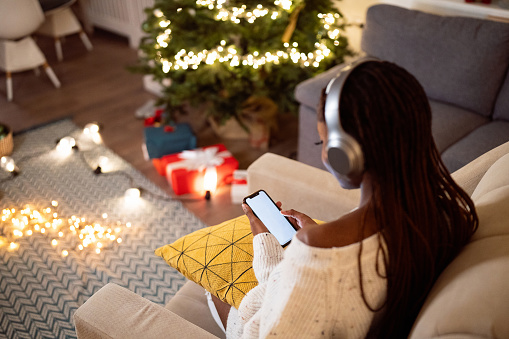 A young woman of Black ethnicity, enjoying a Christmas and New Year atmosphere while listening to music on headphones and using mobile phone