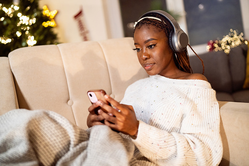 A young woman of Black ethnicity, enjoying a Christmas and New Year atmosphere while listening to music on headphones and using mobile phone