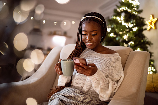 Young woman Black ethnicity, sitting on the arm chair, drinking coffee/tea or hot chocolate and using mobile phone, while enjoying Christmas atmosphere in her home