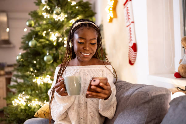 istock Woman Black ethnicity enjoying Christmas atmosphere, while drinking tea and using mobile phone 1875181618