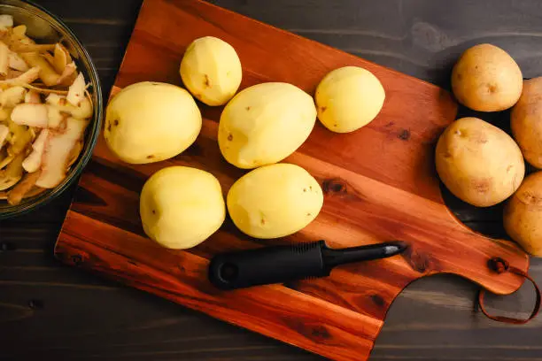 Peeled and unpeeled golden yellow potatoes with a vegetable peeler on a wood table