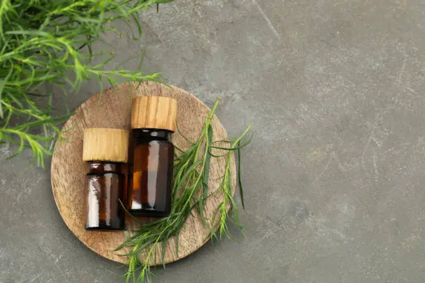 Bottles of essential oil and fresh tarragon leaves on grey table, flat lay. Space for text