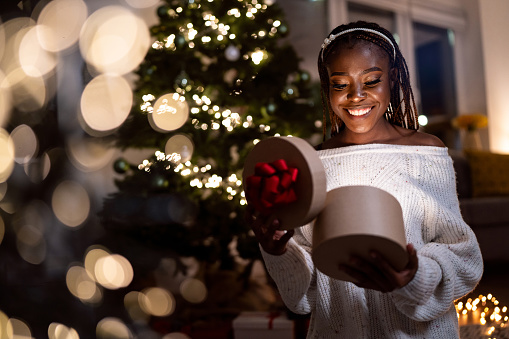 Portrait of an young woman of Black ethnicity sitting neat Christmas tree and opening the present