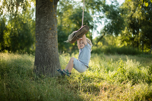 A blond 3 year old boy plays in the village with a swing made of rope and a log. Summer time in the countryside, life in nature.