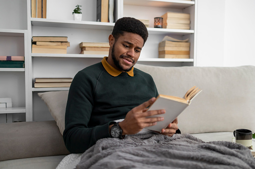 A young American-African man relaxing at home while reading a book in his bed