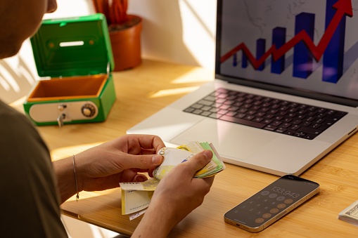 Unrecognizable young Caucasian male freelancer, counting money (euros in 100 and 50 banknotes), while working from the home office