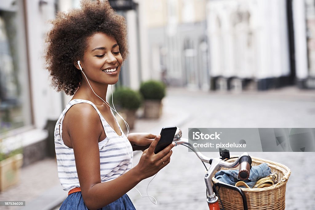 Choosing a song to fit her mood Smiling young woman wearing headphones and pushing her bicycle while checking her cellphone Cycling Stock Photo