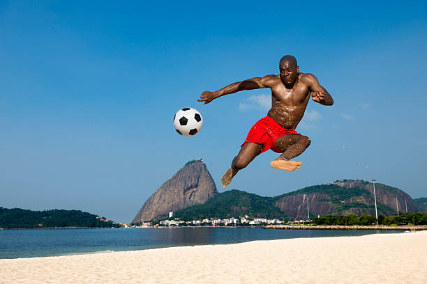 Beach Soccer Player Soccer player kicking the ball in mid air on Flamingo beach, Rio de Janeiro, Brazil. Composite image. brazil beach soccer stock pictures, royalty-free photos & images