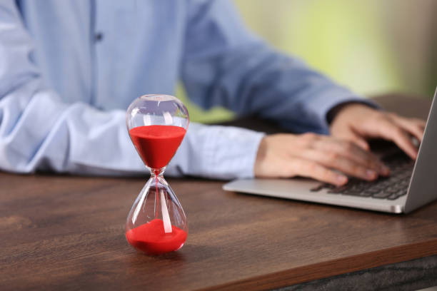istock Hourglass with flowing sand on wooden table, selective focus. Man using laptop indoors 1875415369