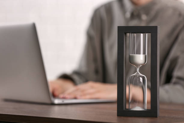 istock Hourglass with flowing sand on wooden table, selective focus. Man using laptop indoors 1875415375
