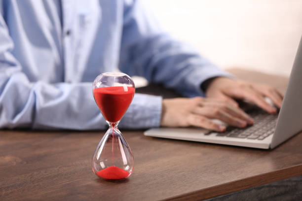 istock Hourglass with flowing sand on wooden table, selective focus. Man using laptop indoors 1875415537