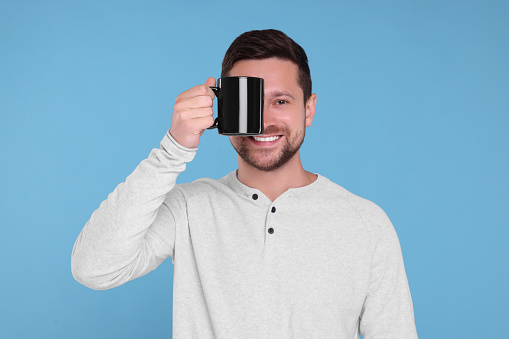 Portrait of happy man covering eye with black mug on light blue background