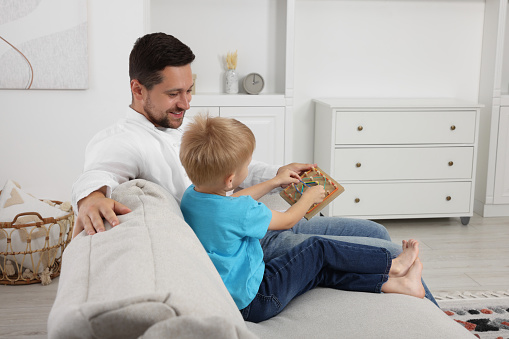 Motor skills development. Happy father helping his son to play with geoboard and rubber bands on sofa at home