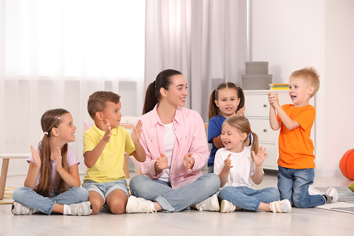 Nursery teacher and group of cute little children spending time together on floor in kindergarten. Playtime activities