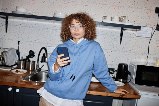 Waist up portrait of curly haired young woman standing in kitchen and looking at camera with flash