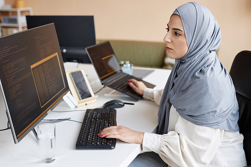Side view portrait of Muslim young woman wearing modest clothing in office while working as female software developer and writing code with multiple computer devices