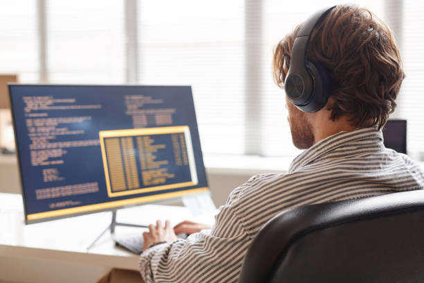 Male IT Developer Using Computer at Desk in Office and Wearing Headphones