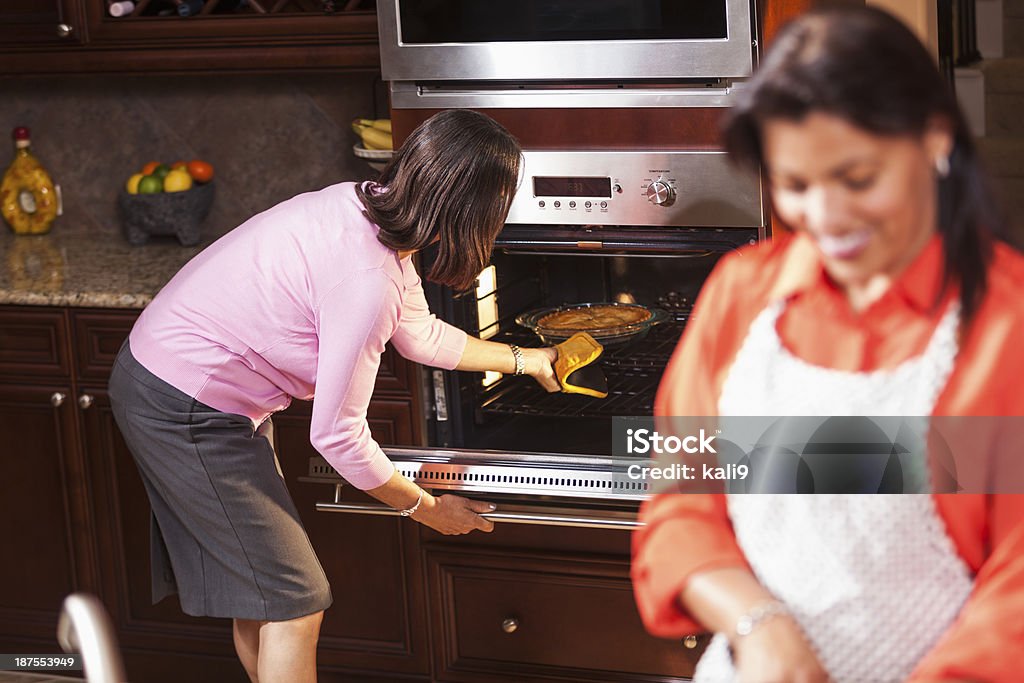 African American women in kitchen cooking dinner. African American women in kitchen preparing food.  Focus on senior woman (60s) checking on pie baking in oven.  Her adult daughter (40s) is standing, smiling in foreground, out of focus. 40-49 Years Stock Photo