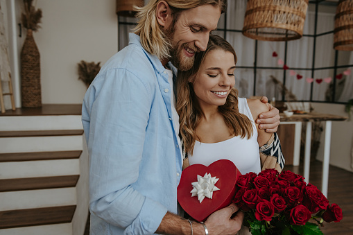 Young loving couple embracing while holding red roses and gift box with decorated living room on background