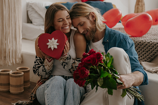 Loving couple embracing and holding roses and gift box while leaning on bed with heart shape balloons on background