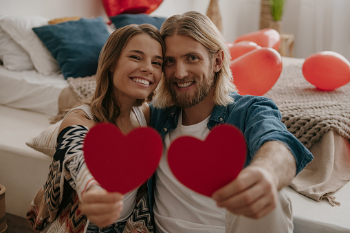 Smiling loving couple embracing and holding heart shape Valentines cards while sitting gin decorated bedroom