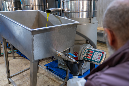 An old Muslim man is filling fresh olive oil into plastic gallons using a funnel and weighing scale in a pressing factory