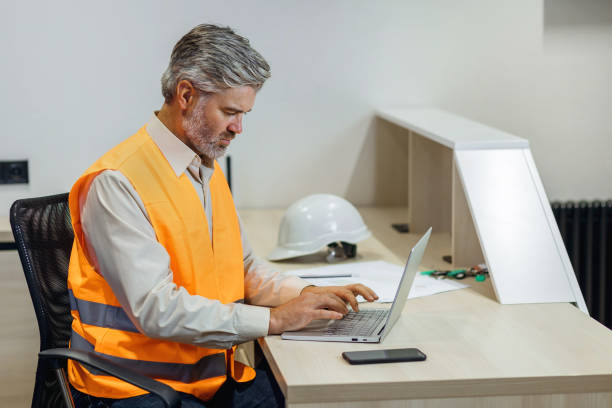 istock gray haired attractive construction bearded man in vest working with laptop, work on Home building project 1875741281