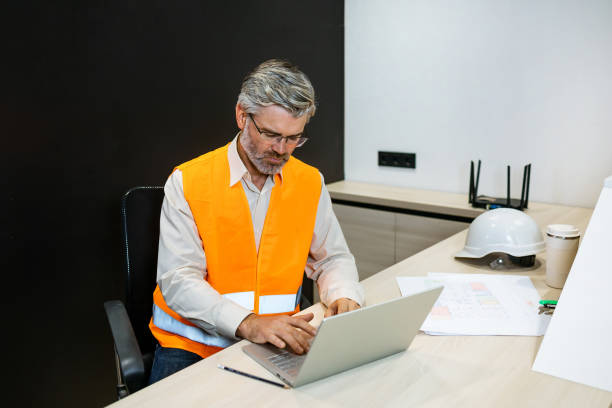 istock senior mature man engineer with laptop computer typing on laptop wear safety vest. Work housing projects and architecture 1875741288