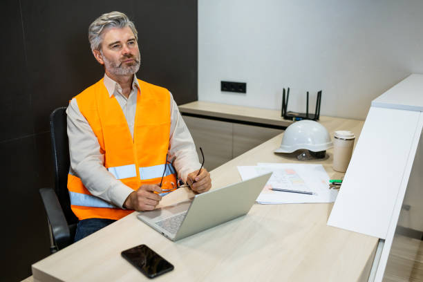 istock Pensive frowning construction engineer sitting at office desk with blueprints and laptop looking away 1875741295