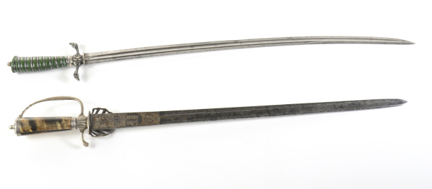 Two late 18th century British swords on a white background.    One a military sword-sabre with a slightly curved blade, double edged near the point, a knuckle guard with twin raptor heads and green and silver wire wrapped handle.  The second is a hunting sword with a sterling hilt dated 1743 with antler/bone shaped handle.