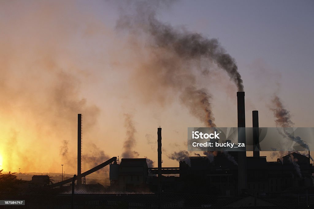 Backlit paper mill factory sunset with dark smoke rising upwards Backlit paper mill at sunset with dark smoke rising upwards into the orange sky Greenhouse Stock Photo